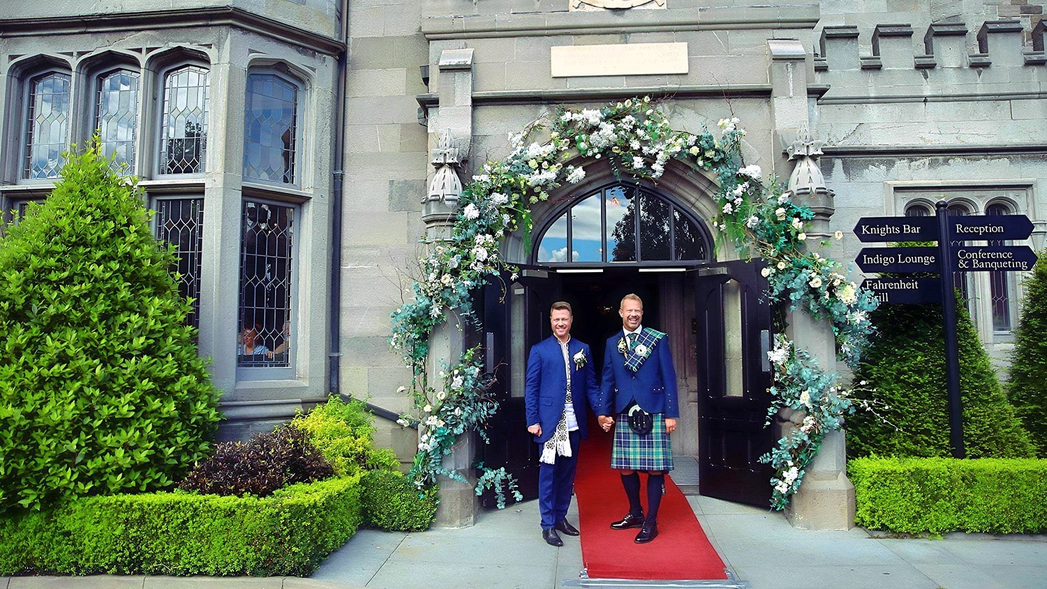 A man in a suit and another in a kilt stand hand in hand at the exterior of Clontarf Castle, the stand on a red carpet as they enter into a civil ceremony