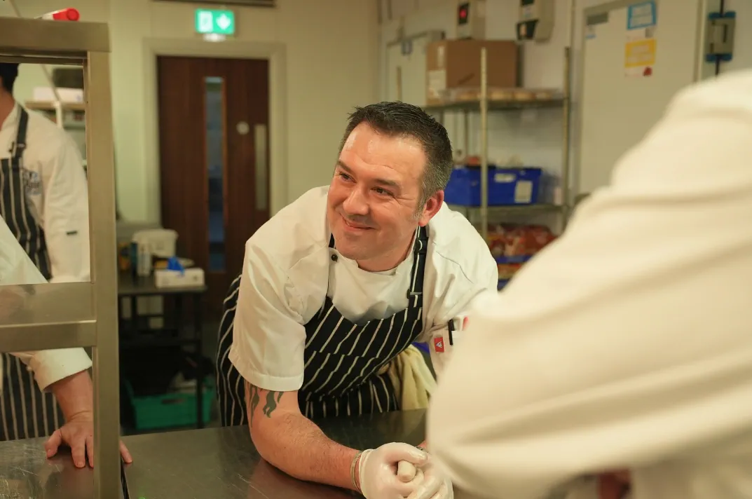 A portrait head shot of Executive Head Chef Paul Devoy. He is smiling looking straight to the camera, contrasting from a grey wall behind him.